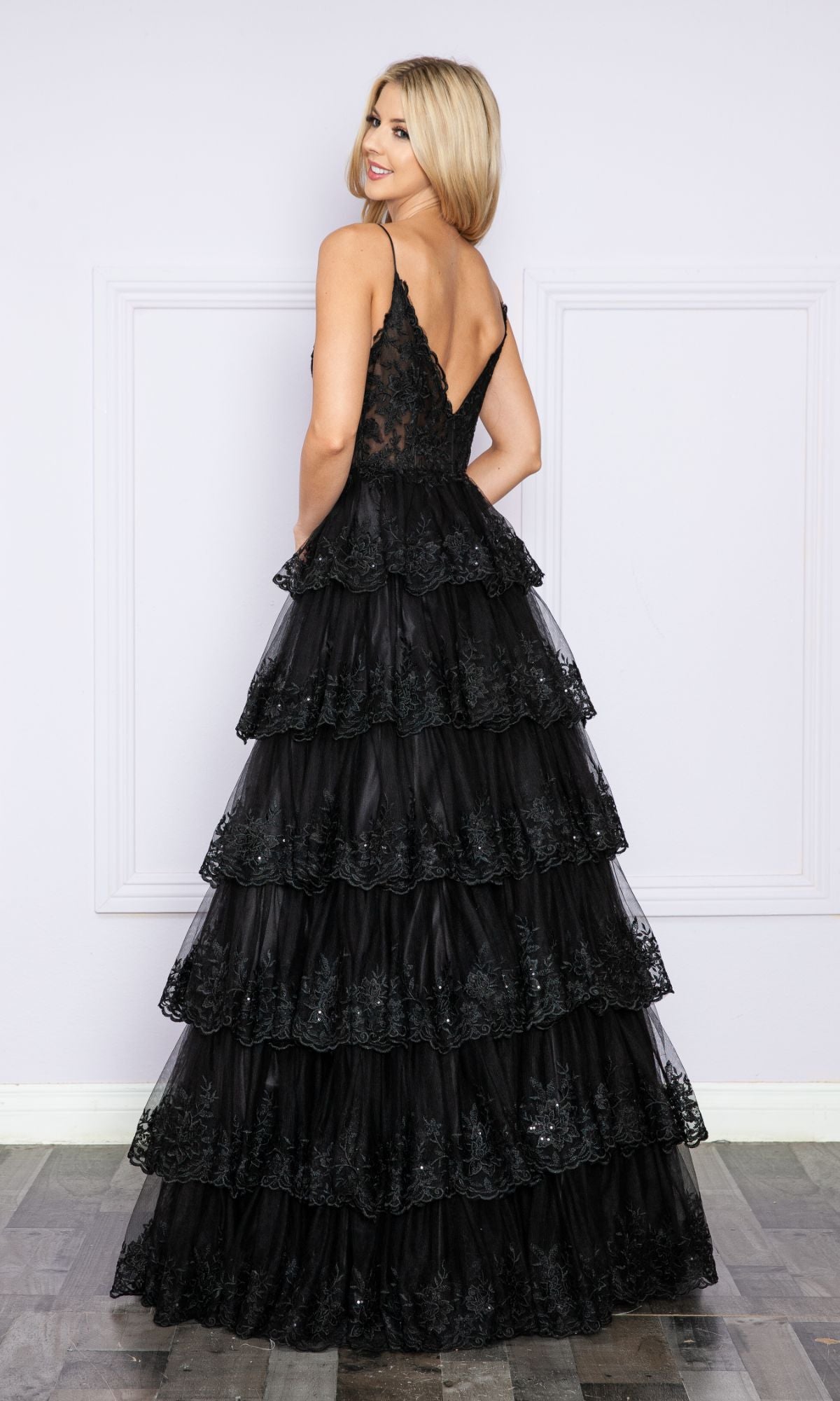 A woman stands facing away in a Sheer-Bodice Long Ruffle Prom Ball Gown: Poly 9402, showcasing its sleeveless design, embroidered-lace bodice, deep V-back, and layered ruffles in a room with light walls and wood floors.